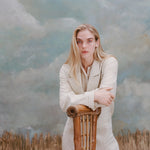 Female model with long-blonde hair with crossed arms leaning on a bamboo chair wearing the 'Easy-Going Jacket' in sand linen with a beige collared shirt, shot on a light blue textured background.