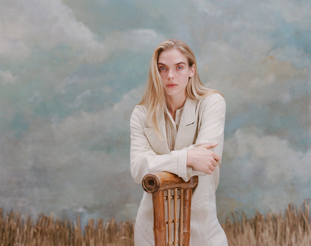 Female model with long-blonde hair with crossed arms leaning on a bamboo chair wearing the 'Easy-Going Jacket' in sand linen with a beige collared shirt, shot on a light blue textured background.