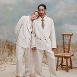 Male model with dreadlock and female model with long-blonde hair both wearing the 'Easy-Going Jacket' in sand linen with matching sand linen pants, and beige shoes, shot on a light blue textured background with waist-high beige grass and a bamboo chair.
