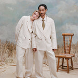 Male model with dreadlock and female model with long-blonde hair both wearing the 'Easy-Going Jacket' in sand linen with matching sand linen pants, and beige shoes, shot on a light blue textured background with waist-high beige grass and a bamboo chair.