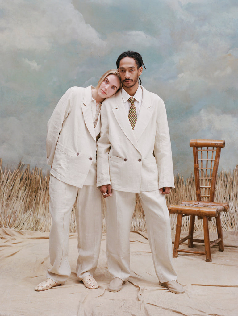 Male model with dreadlock and female model with long-blonde hair both wearing the 'Easy-Going Jacket' in sand linen with matching sand linen pants, and beige shoes, shot on a light blue textured background with waist-high beige grass and a bamboo chair.