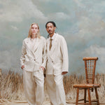 Male model with dreadlocks and female model with long blonde hair standing next to one another wearing the 'Easy-Going Pants' in Sand Linen with matching blazers in sand linen, with the female wearing an beige unbuttoned collared shirt with beige loafers and the male wearing a beige button down shirt and brown with yellow patterned tie with beige slip-ons, shot on a light-blue textured background with waist-high beige sand and a bamboo chair also in the background.