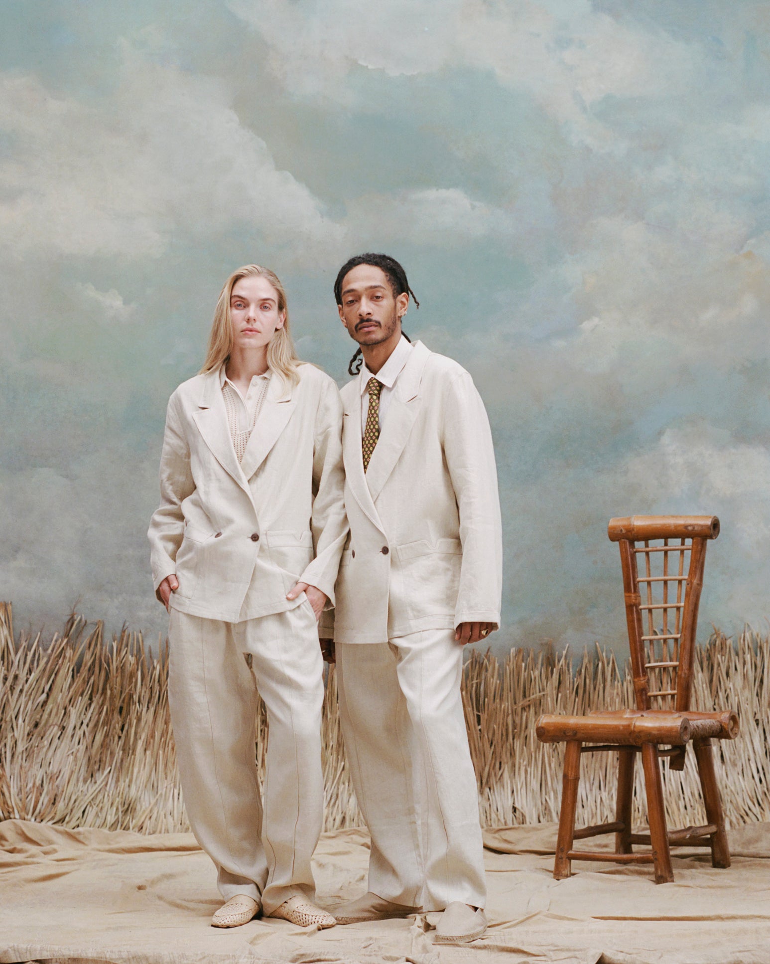 Male model with dreadlocks and female model with long blonde hair standing next to one another wearing the 'Easy-Going Pants' in Sand Linen with matching blazers in sand linen, with the female wearing an beige unbuttoned collared shirt with beige loafers and the male wearing a beige button down shirt and brown with yellow patterned tie with beige slip-ons, shot on a light-blue textured background with waist-high beige sand and a bamboo chair also in the background.