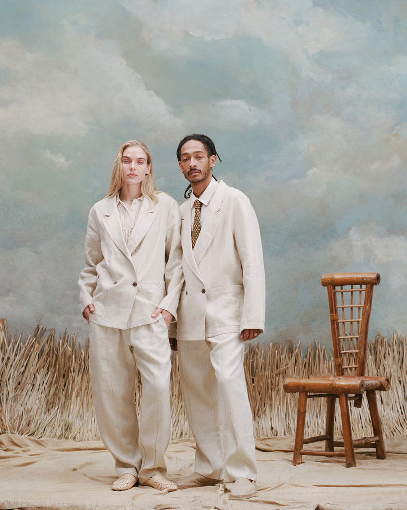 Male model with dreadlocks and female model with long blonde hair standing next to one another wearing the 'Easy-Going Pants' in Sand Linen with matching blazers in sand linen, with the female wearing an beige unbuttoned collared shirt with beige loafers and the male wearing a beige button down shirt and brown with yellow patterned tie with beige slip-ons, shot on a light-blue textured background with waist-high beige sand and a bamboo chair also in the background.
