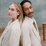 Female model with long-blonde hair and male model with dreadlocks standing back to back both wearing the 'Easy-Going Jacket' in sand linen while the male model wears a green tie with red and yellow dice patterning, shot on a light blue textured background.