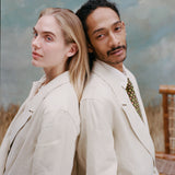 Female model with long-blonde hair and male model with dreadlocks standing back to back both wearing the 'Easy-Going Jacket' in sand linen while the male model wears a green tie with red and yellow dice patterning, shot on a light blue textured background.