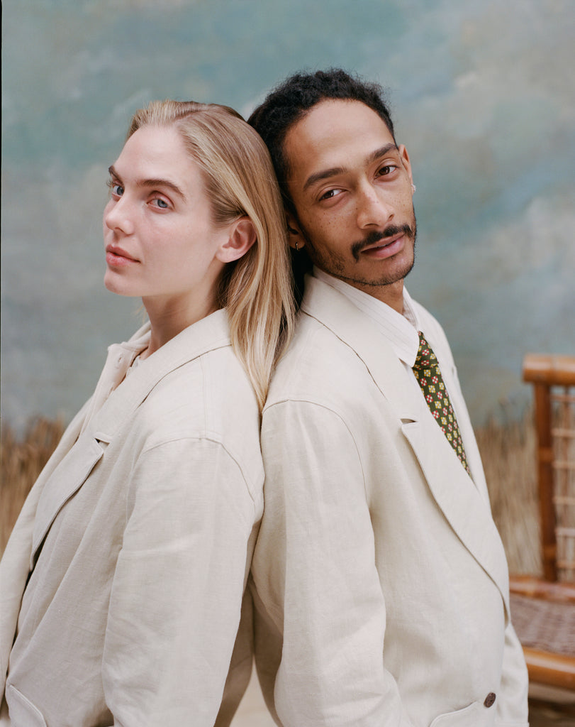 Female model with long-blonde hair and male model with dreadlocks standing back to back both wearing the 'Easy-Going Jacket' in sand linen while the male model wears a green tie with red and yellow dice patterning, shot on a light blue textured background.