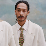 Male model with dreadlocks wearing the 'Easy-Going Jacket' in sand linen with a collared white shirt and green tie with red and yellow dice.