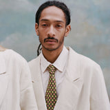 Male model with dreadlocks wearing the 'Easy-Going Jacket' in sand linen with a collared white shirt and green tie with red and yellow dice.