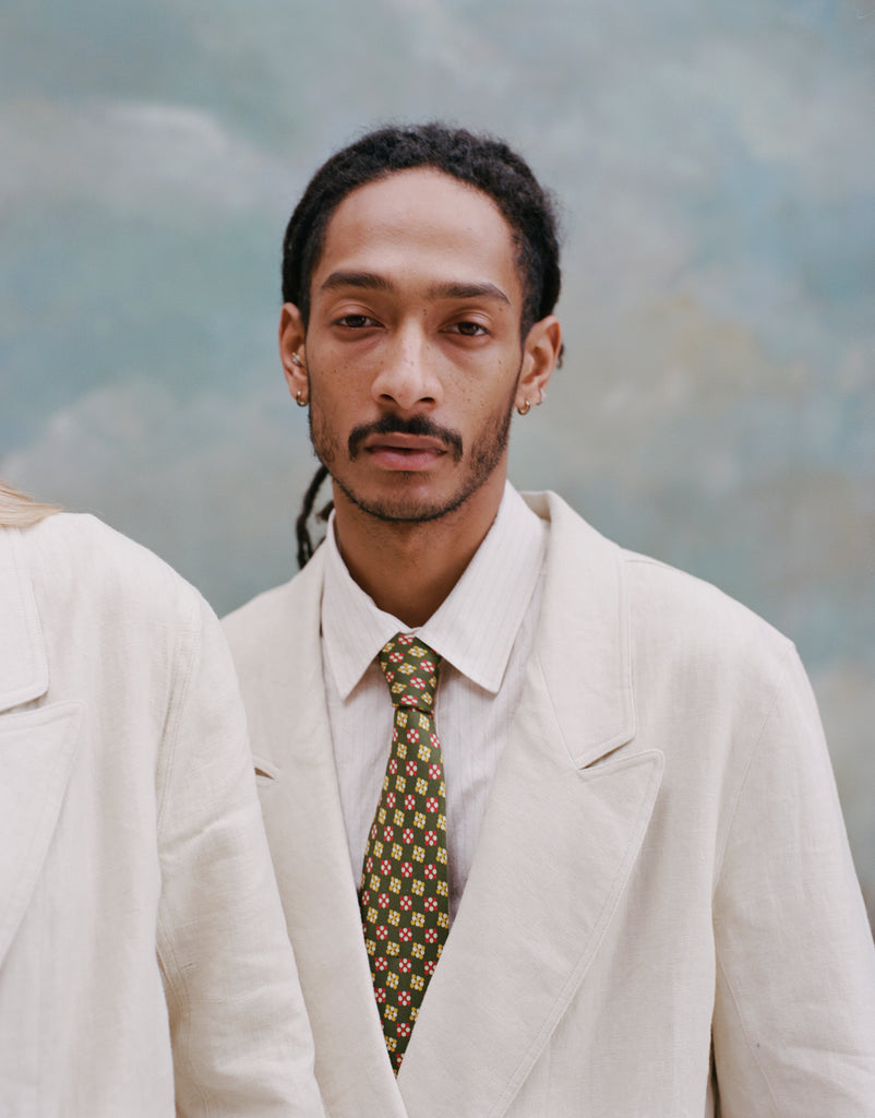 Male model with dreadlocks wearing the 'Easy-Going Jacket' in sand linen with a collared white shirt and green tie with red and yellow dice.