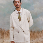 Male model wearing the 'Easy-Going Jacket' in sand linen with a white collared shirt and brown/yellow pattern tie and matching sand linen pants, shot on a light-blue textured background with waist high beige grass.