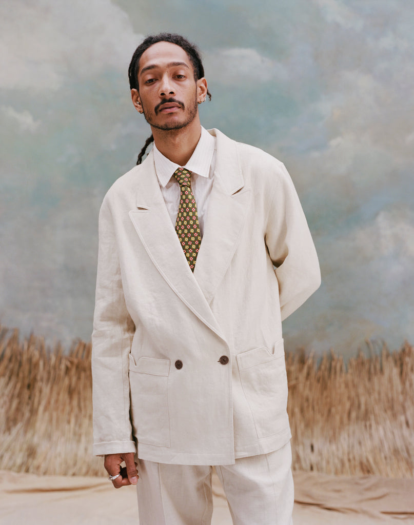Male model wearing the 'Easy-Going Jacket' in sand linen with a white collared shirt and brown/yellow pattern tie and matching sand linen pants, shot on a light-blue textured background with waist high beige grass.