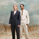 Female model with long blonde hair wearing the 'easy-going' jacket in navy jacquard stripe with matching pants and beige loafers while standing beside a male model with dreadlocks wearing an 'easy-going' jacket in white with matching patns, a white collared shirt and brown and yellow tie underneath, with white slippers, shot on a light blue textured background with beige grass.