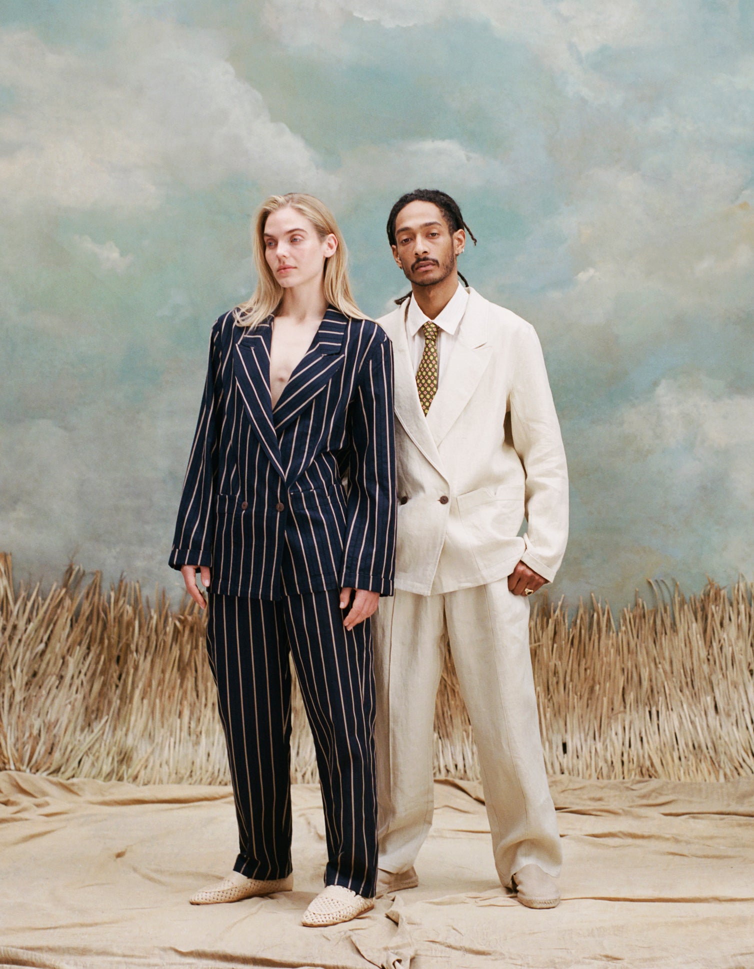 Female model with long blonde hair wearing the 'easy-going' jacket in navy jacquard stripe with matching pants and beige loafers while standing beside a male model with dreadlocks wearing an 'easy-going' jacket in white with matching patns, a white collared shirt and brown and yellow tie underneath, with white slippers, shot on a light blue textured background with beige grass.