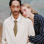 Male model with dreadlocks and female model with long blond hair resting her head on the male model with her arms wrapped around his, while he wears the 'Easy-Going' jacket in sand linen with a white collared shirt and green tie with dice patterning, while the female model wears a navy and beige striped blazer, shot a light blue textured background.