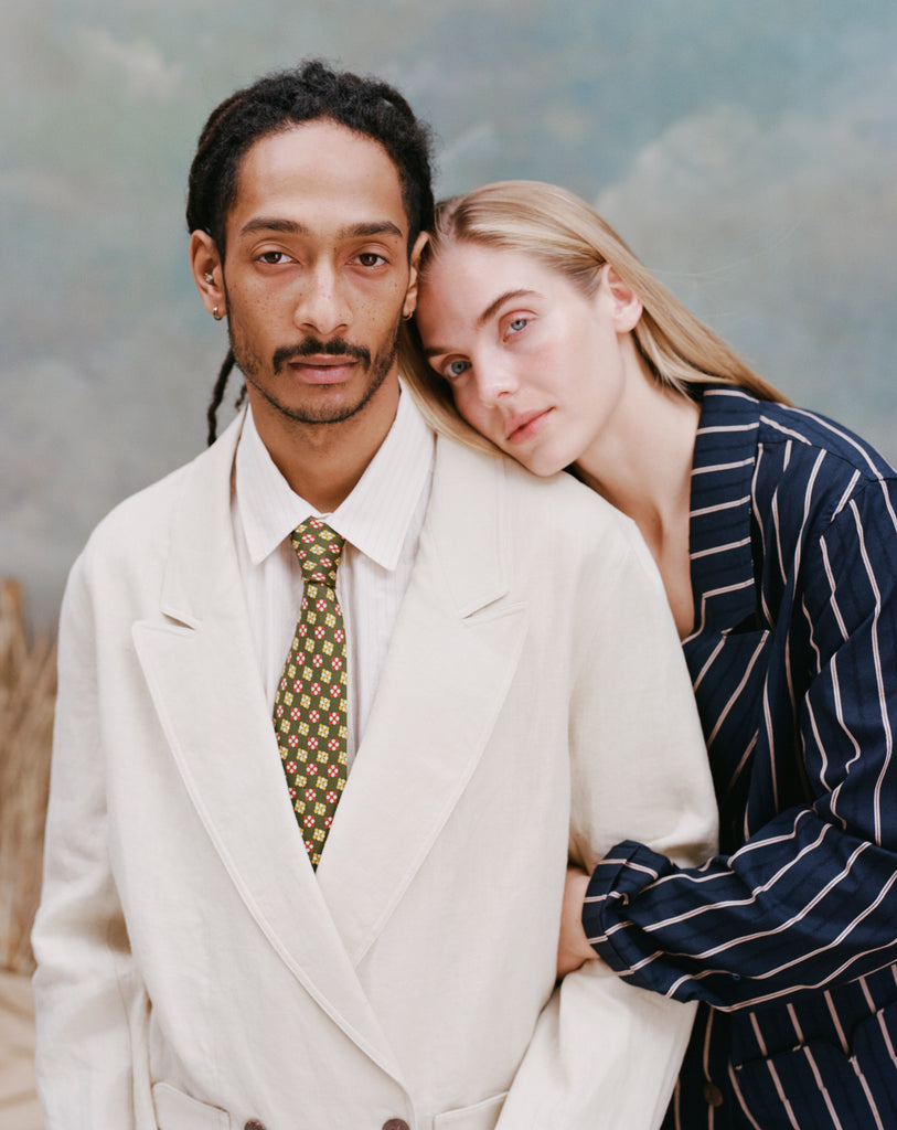 Male model with dreadlocks and female model with long blond hair resting her head on the male model with her arms wrapped around his, while he wears the 'Easy-Going' jacket in sand linen with a white collared shirt and green tie with dice patterning, while the female model wears a navy and beige striped blazer, shot a light blue textured background.
