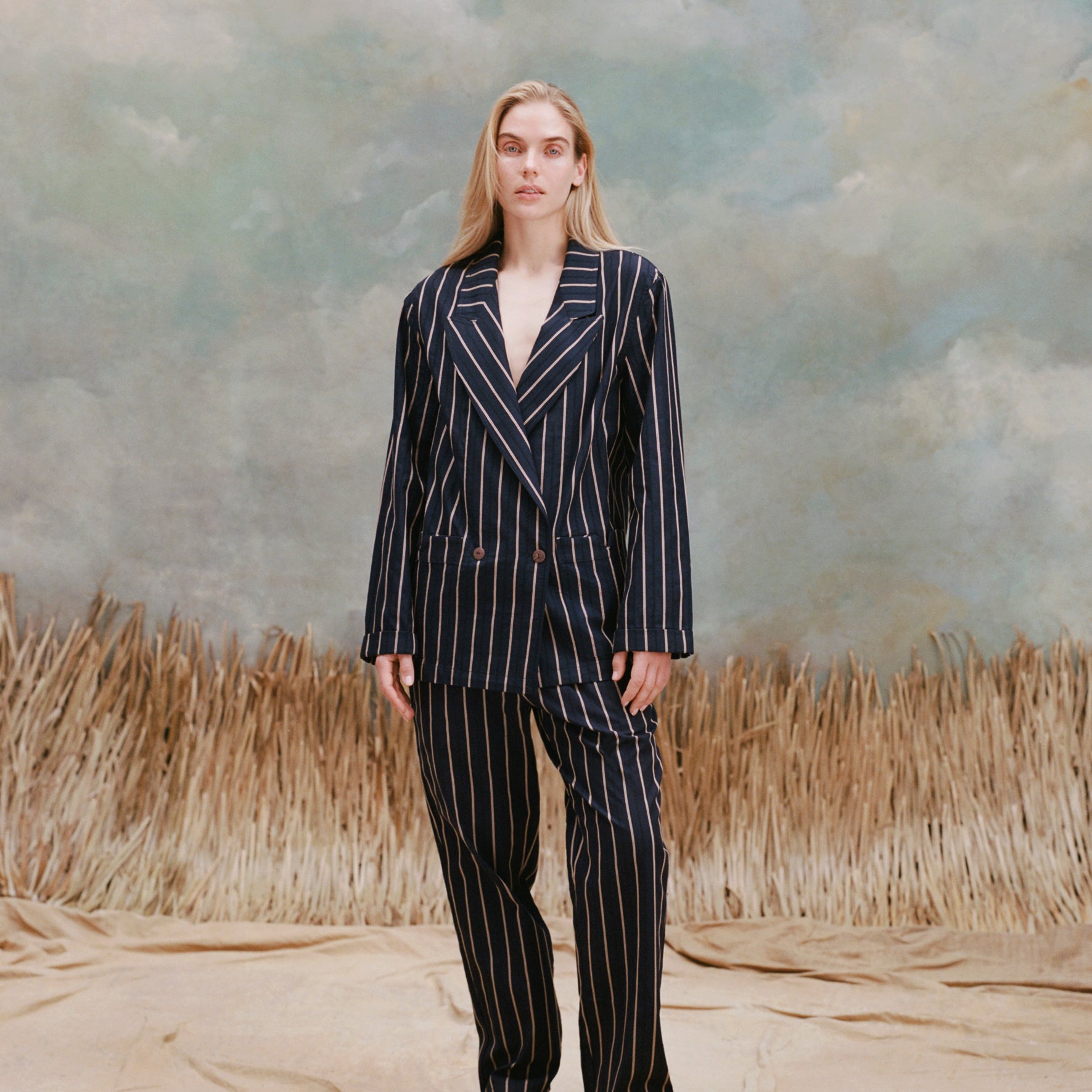 Female model with long-blonde hair wearing the 'Easy-Going Pants' in a navy jacquard stripe with a matching navy jacquard stripe blazer, and brown loafers, shot on a light-blue textured background, and waist-high beige grass.