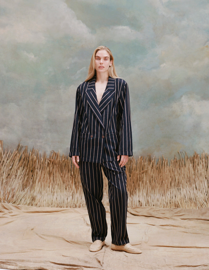 Female model with long-blonde hair wearing the 'Easy-Going Pants' in a navy jacquard stripe with a matching navy jacquard stripe blazer, and brown loafers, shot on a light-blue textured background, and waist-high beige grass.