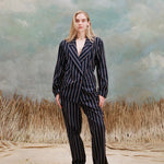 Female model with long-blonde hair wearing the 'easy-going jacket' in navy jacquard stripe with matching navy jacquard striped pants and beige loafers, shot on a light blue textured background with beige grass.