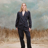 Female model with long-blonde hair wearing the 'easy-going jacket' in navy jacquard stripe with matching navy jacquard striped pants and beige loafers, shot on a light blue textured background with beige grass.
