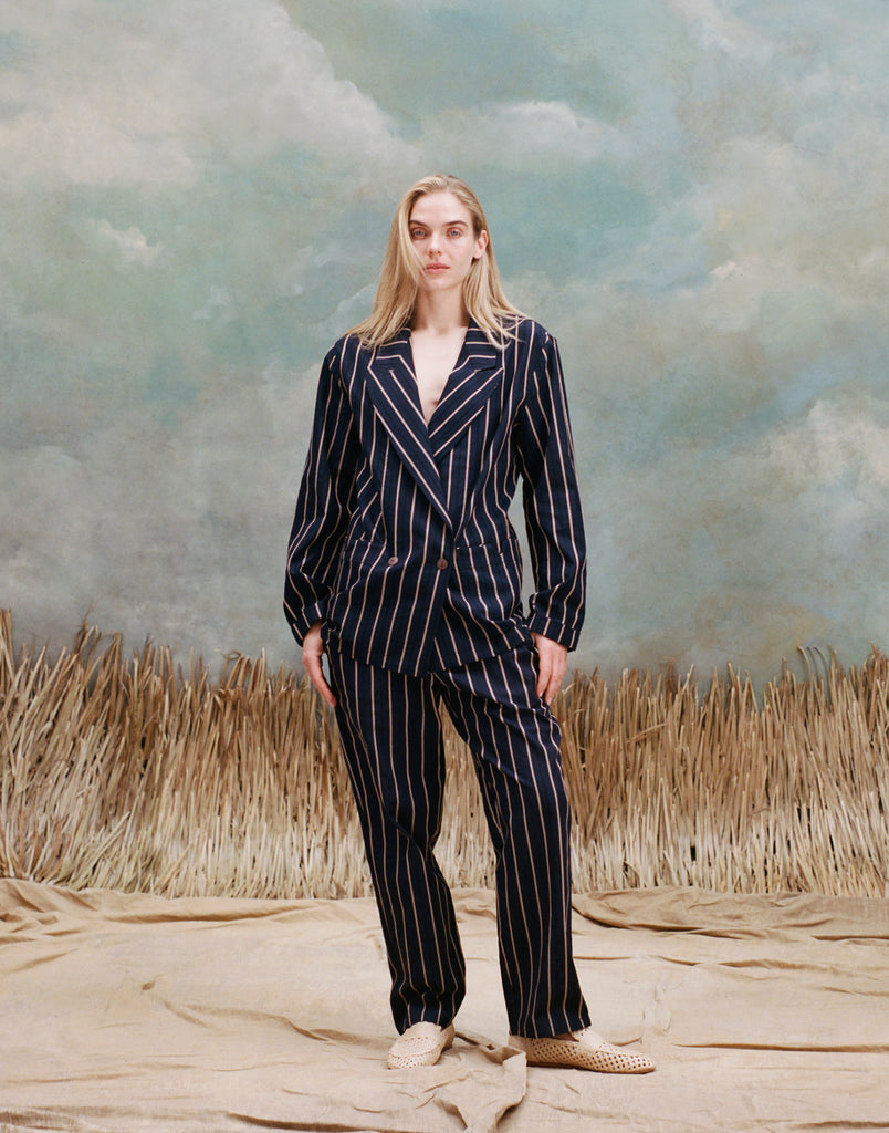 Female model with long-blonde hair wearing the 'easy-going jacket' in navy jacquard stripe with matching navy jacquard striped pants and beige loafers, shot on a light blue textured background with beige grass.