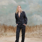 Female model with long-blonde hair wearing the 'easy-going' jacket in navy jacquard stripe with matching navy jacquard pants, and beige loafers, shot on a light blue textured background with beige grass.