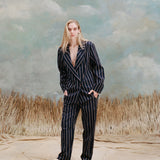 Female model with long-blonde hair wearing the 'easy-going' jacket in navy jacquard stripe with matching navy jacquard pants, and beige loafers, shot on a light blue textured background with beige grass.
