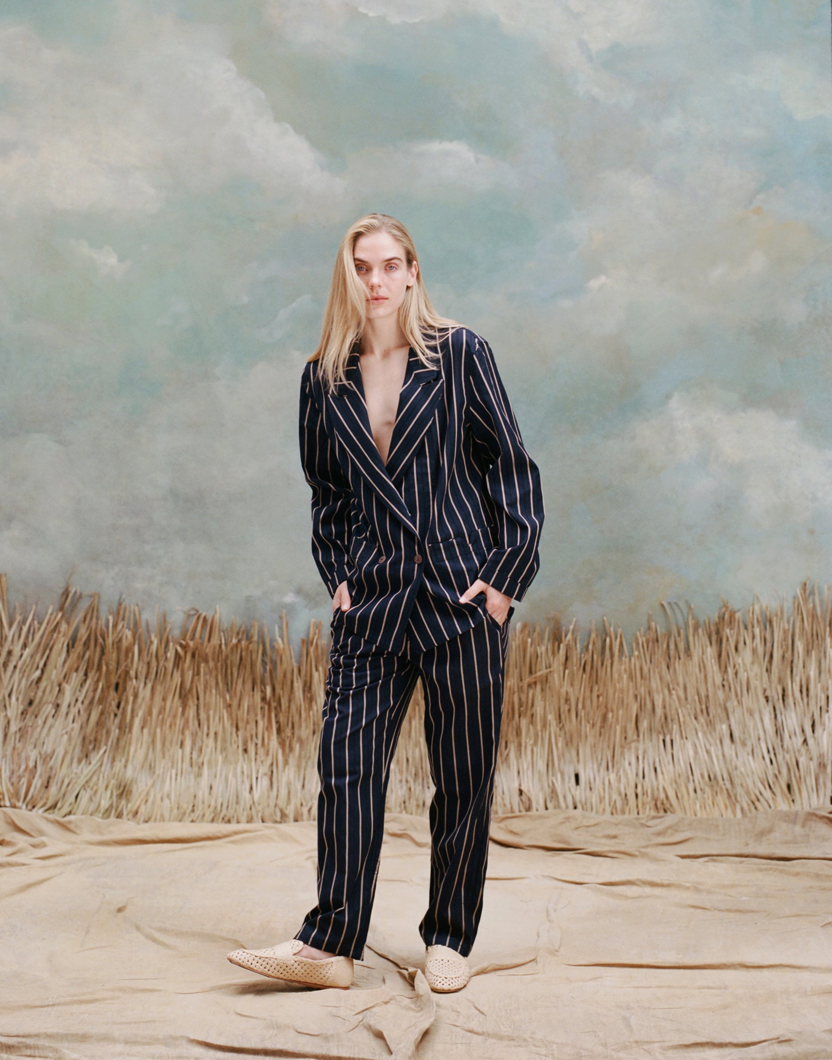 Female model with long-blonde hair wearing the 'easy-going' jacket in navy jacquard stripe with matching navy jacquard pants, and beige loafers, shot on a light blue textured background with beige grass.