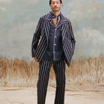 Male model with dreadlocks wearing the 'easy-going' jacket in navy jacquard stripe with matching navy-jacquard striped pants with navy espadrills and a navy cabana shirt shot on a light blue textured background with beige grass.