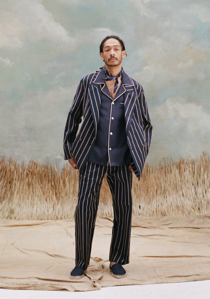 Male model with dreadlocks wearing the 'Easy-Going Pants' in Navy Jacquard Stripe and a matching Navy Jacquard blazer with a navy button-down shirt with brown trim and a navy handkerchief around his neck, with a navy slip-on shoes, shot on a light-blue textured background, with waist-high beige grass.