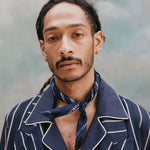 Male model with dreadlocks wearing th e'easy-going' jacket in navy jacquard stripe with a navy cabana shirt underneath and navy and white patterned cloth around his neck, shot on a light blue textured background.