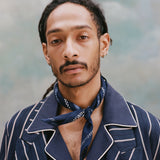 Male model with dreadlocks wearing th e'easy-going' jacket in navy jacquard stripe with a navy cabana shirt underneath and navy and white patterned cloth around his neck, shot on a light blue textured background.