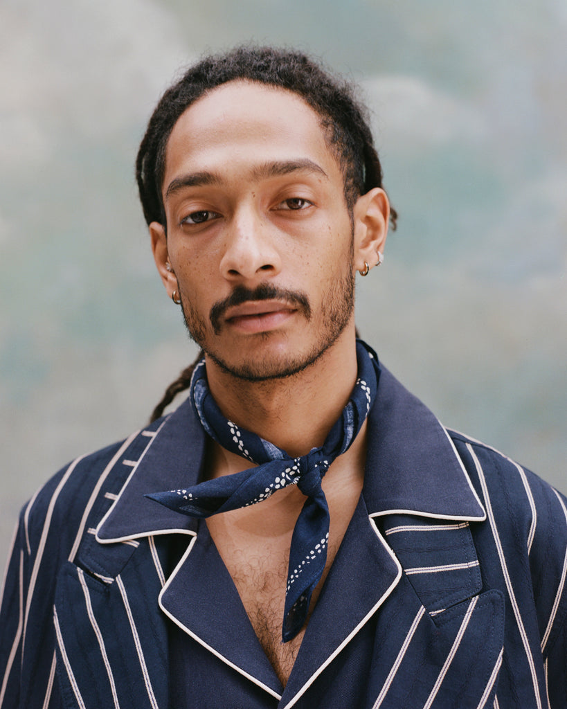 Male model with dreadlocks wearing th e'easy-going' jacket in navy jacquard stripe with a navy cabana shirt underneath and navy and white patterned cloth around his neck, shot on a light blue textured background.