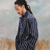 Male model with dreadlocks wearing the 'easy-going' jacket in navy jacquard stripe with a navy cabana shirt underneath and a navy and white patterned cloth wrapped around his neck, shot on a light blue textured background with beige grass.