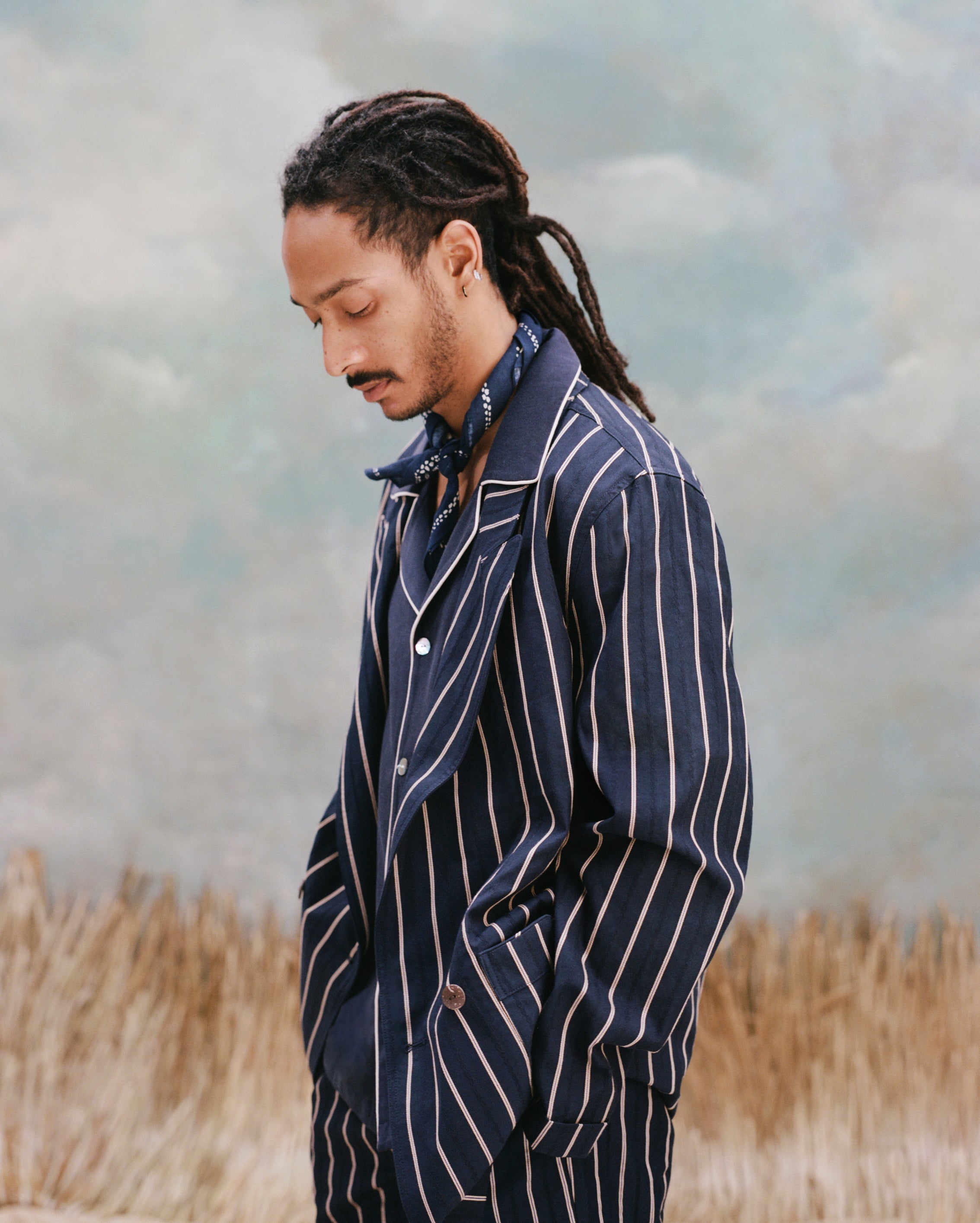 Male model with dreadlocks wearing the 'easy-going' jacket in navy jacquard stripe with a navy cabana shirt underneath and a navy and white patterned cloth wrapped around his neck, shot on a light blue textured background with beige grass.