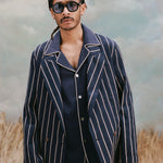 Male model with dreadlocks wearing the 'easy-going' jacket in navy jacquard stripes with a navy cabana shirt underneath and a navy jacquard stripe pants, and black rounded-square sunglasses with light blue tinted lenses, shot on a light blue textured background with beige grass.