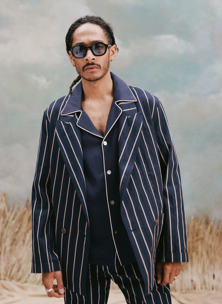 Male model with dreadlocks wearing the 'easy-going' jacket in navy jacquard stripes with a navy cabana shirt underneath and a navy jacquard stripe pants, and black rounded-square sunglasses with light blue tinted lenses, shot on a light blue textured background with beige grass.