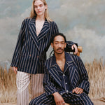Male model with dreadlocks seated wearing the 'easy-going' jacket in navy jacquard stripe with matching navy jacquard striped pants, and a navy shirt underneath, and a female model with long-blonde hair wearing the 'easy-going' jacket in navy jacquard stripe with white and brown stripe pants, shot on a light blue textured background with beige grass.
