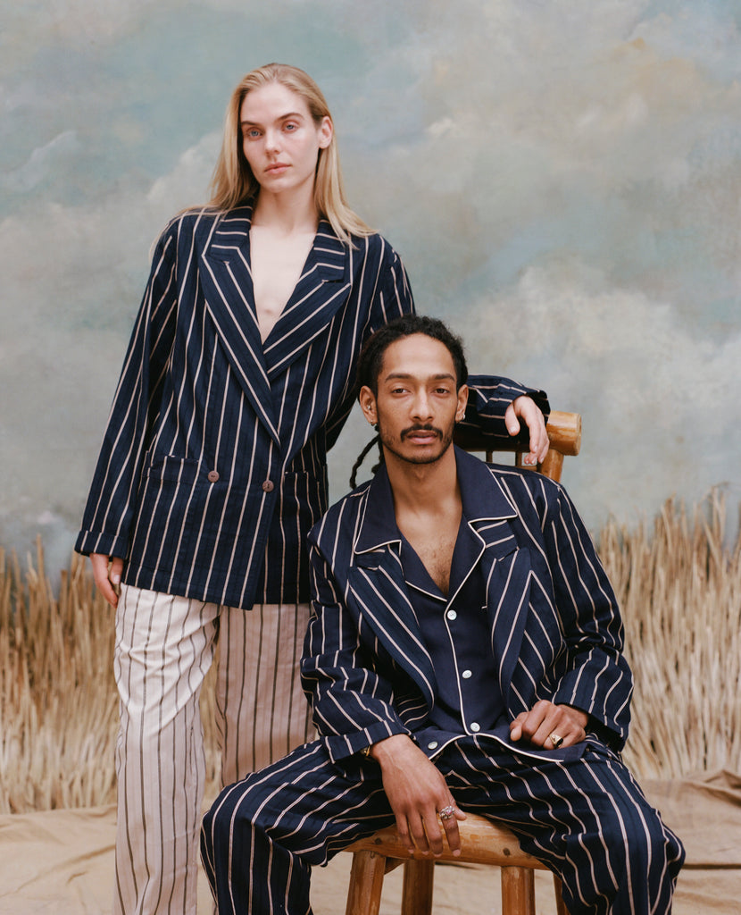 Male model with dreadlocks seated wearing the 'easy-going' jacket in navy jacquard stripe with matching navy jacquard striped pants, and a navy shirt underneath, and a female model with long-blonde hair wearing the 'easy-going' jacket in navy jacquard stripe with white and brown stripe pants, shot on a light blue textured background with beige grass.