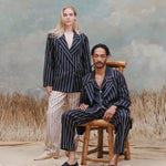 Female model with long blonde hair wearing the 'easy-going jacket' in navy jacquard stripe with matching navy jacquard pants, and beige loafers, standing behind a male model who is seated wearing the 'easy-going' jacket in navy jacquard stripe with a navy cabana shirt underneath and a matching navy jacquard pants with navy espadrilles, shot on a light blue textured background with beige grass.