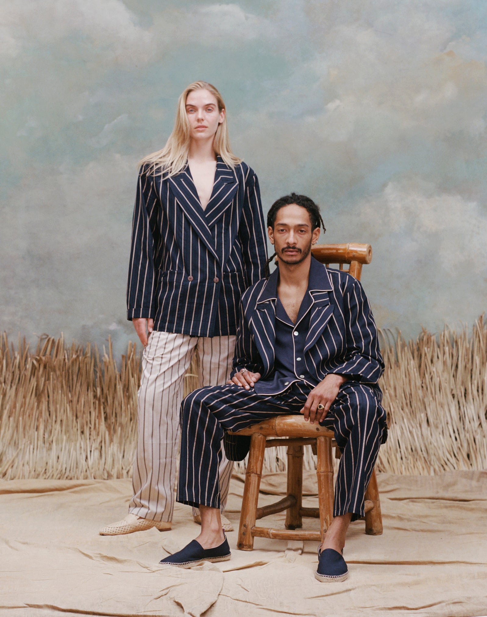 Female model with long blonde hair wearing the 'easy-going jacket' in navy jacquard stripe with matching navy jacquard pants, and beige loafers, standing behind a male model who is seated wearing the 'easy-going' jacket in navy jacquard stripe with a navy cabana shirt underneath and a matching navy jacquard pants with navy espadrilles, shot on a light blue textured background with beige grass.