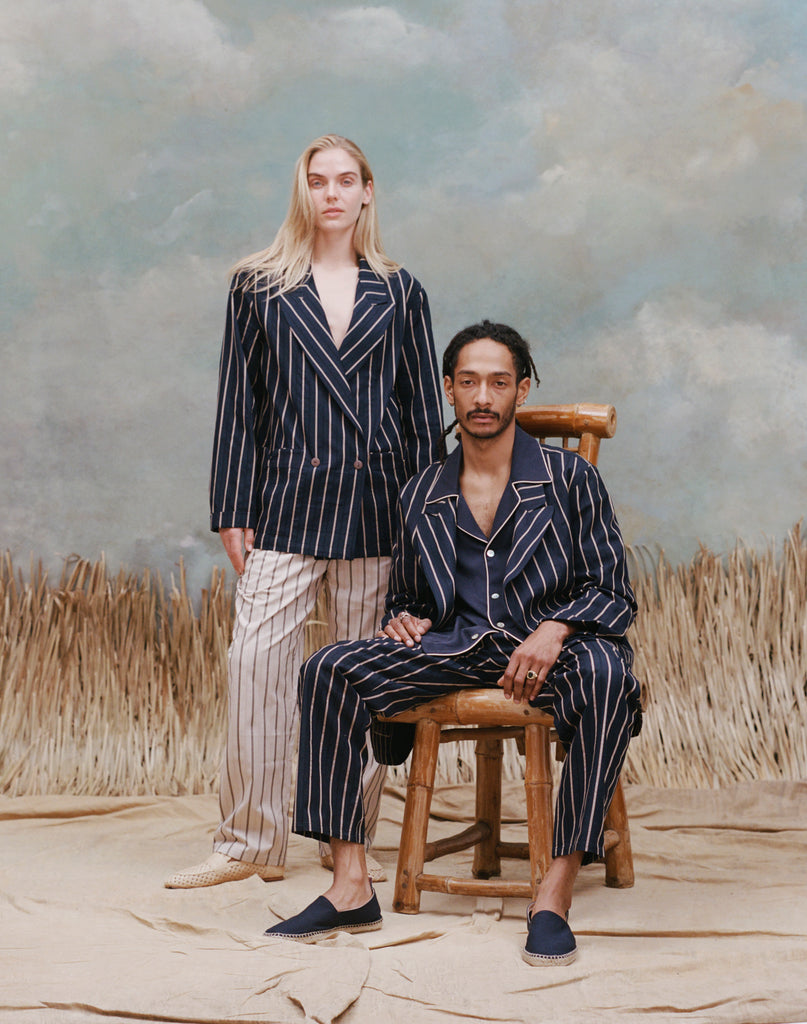 Female model with long blonde hair wearing the 'easy-going jacket' in navy jacquard stripe with matching navy jacquard pants, and beige loafers, standing behind a male model who is seated wearing the 'easy-going' jacket in navy jacquard stripe with a navy cabana shirt underneath and a matching navy jacquard pants with navy espadrilles, shot on a light blue textured background with beige grass.