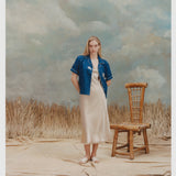 Female model with long blonde hair wearing the 'Teamwork' Cabana ~ Crop Cut unbuttoned with a gold-colored full-length silk dress and white sandals against a background of light blue and white textured paint, waist high beige grass, and a bamboo chair.