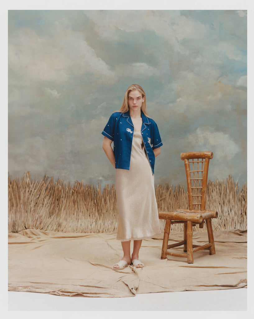 Female model with long blonde hair wearing the 'Teamwork' Cabana ~ Crop Cut unbuttoned with a gold-colored full-length silk dress and white sandals against a background of light blue and white textured paint, waist high beige grass, and a bamboo chair.