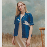 Female model with long blonde hair wearing the 'Teamwork' Cabana ~ Crop Cut unbuttoned with a gold colored silk body length dress against a background of textured ligth blue and white coloring, brown waist high grass, and a bamboo chair.