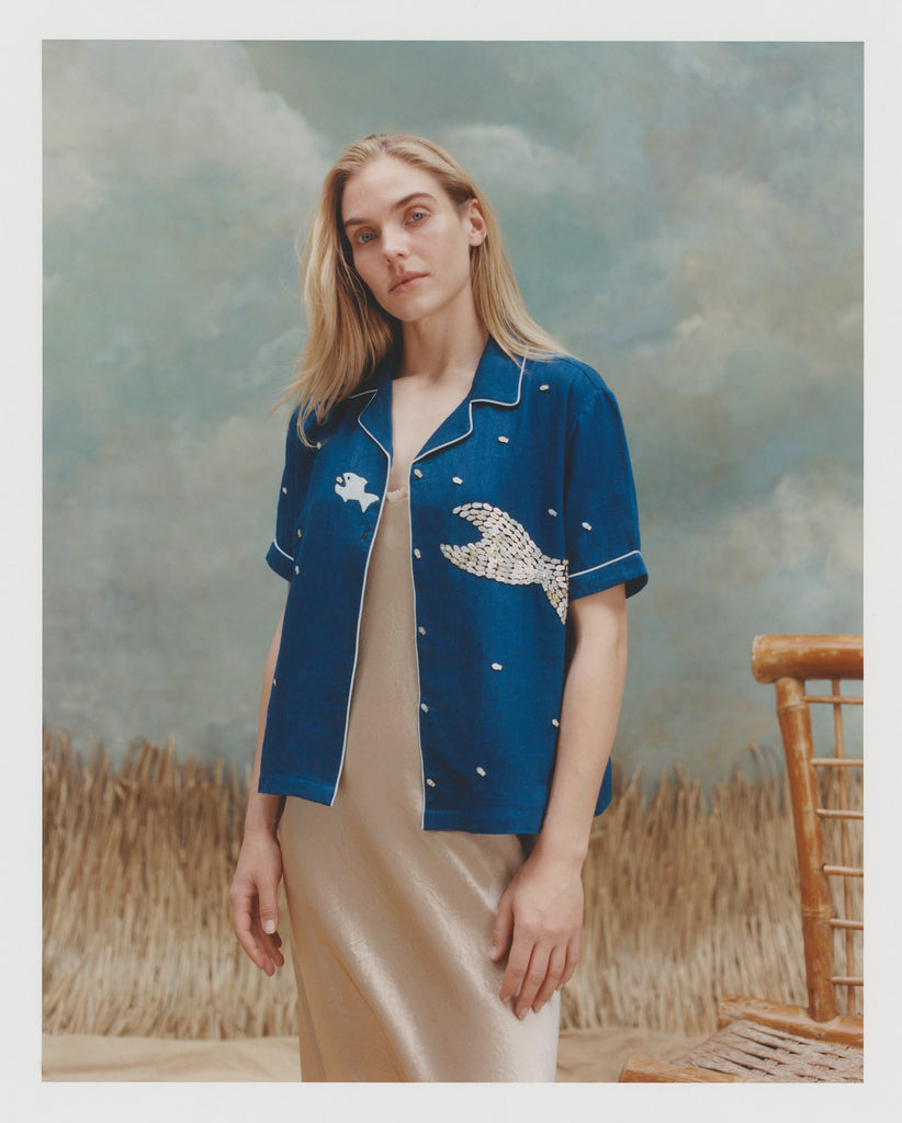 Female model with long blonde hair wearing the 'Teamwork' Cabana ~ Crop Cut unbuttoned with a gold colored silk body length dress against a background of textured ligth blue and white coloring, brown waist high grass, and a bamboo chair.