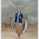 Female model with long blonde hair wearing an unbuttoned 'Teamwork' Cabana ~ Crop Cut with a full-length gold-colored silk dress and white sandals while holding two replica model fish against a light blue and white textured background with waist high beige grass.