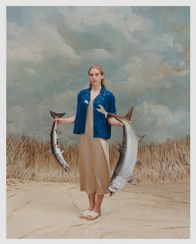 Female model with long blonde hair wearing an unbuttoned 'Teamwork' Cabana ~ Crop Cut with a full-length gold-colored silk dress and white sandals while holding two replica model fish against a light blue and white textured background with waist high beige grass.