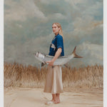Female model with long blonde hair wearing the 'Teamwork' Cabana ~ Crop Cut while holding a model fish, and wearing a gold colored silk dress and white sandals, all shot on a background of texutred light blue and white with waist high beige grass.