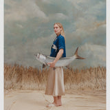 Female model with long blonde hair wearing the 'Teamwork' Cabana ~ Crop Cut while holding a model fish, and wearing a gold colored silk dress and white sandals, all shot on a background of texutred light blue and white with waist high beige grass.
